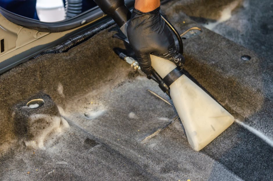 A professional cleaning technician wearing a black glove is using a portable steam or hot water extraction machine to deep clean a dark grey carpeted surface in an indoor room, likely a commercial or
