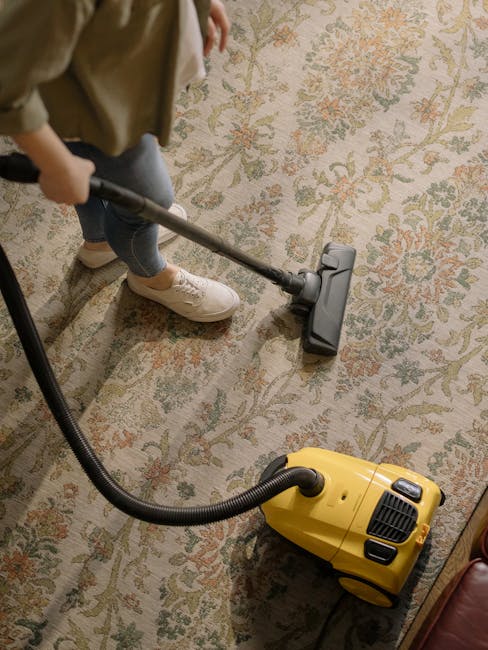 A person using a handheld vacuum cleaner to clean a patterned area rug with floral motifs in a residential room. The individual is dressed in casual attire, including beige shoes and blue jeans, stand